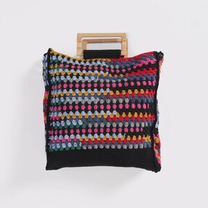 Peut inclure: Un sac fourre-tout au crochet multicolore avec une poignée en bois. Le sac présente une base et une bordure noires, avec un motif de différentes couleurs, dont le rouge, le jaune, le bleu et le rose. La poignée est en bois clair.