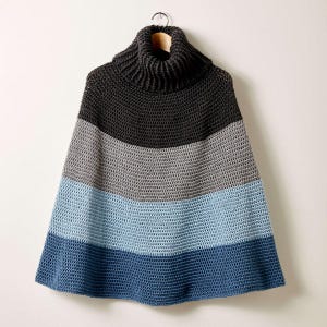 May include: A handmade, crocheted poncho with a turtleneck collar. The poncho features horizontal stripes in shades of charcoal, gray, light blue, and navy blue. The garment is hanging on a wooden hanger against a white background.