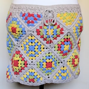 May include: A crocheted skirt featuring a patchwork design with colorful squares in red, yellow, blue, and green. The skirt has a neutral-colored waistband with a drawstring closure. The skirt is made of yarn.