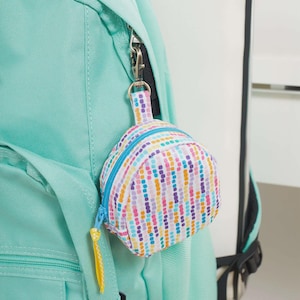 Puede incluir: Un pequeño monedero con forma de mochila colorida con cremallera y clip, sujeto a una mochila turquesa. El monedero presenta un patrón de líneas verticales en varios colores, incluyendo rosa, morado, naranja y azul.
