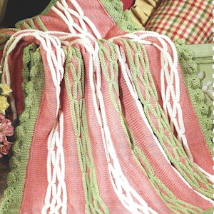 May include: A knitted blanket in shades of pink, white, and green. The blanket features a cable knit design with white and green vertical stripes. The edges are trimmed with a green scalloped border. Pillows in the background add to the cozy aesthetic.