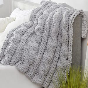 May include: A light gray, chunky knit blanket with a cable-knit design. The blanket is draped over a bed, showcasing its thick, textured weave. The soft, plush material suggests warmth and comfort.