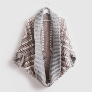 Pode incluir: Um cardigan de crochê feito à mão, com frente aberta, com um padrão de quadrados da vovó. A paleta de cores inclui tons de cinza, bege e malva. O cardigan apresenta uma gola larga e canelada e é exibido em um cabide branco.