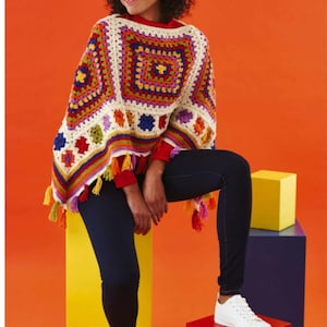 May include: A colorful, crocheted poncho with a square pattern in shades of red, orange, yellow, green, blue, and purple. The poncho has a cream-colored base and is trimmed with tassels in various colors. The person is wearing dark blue jeans and white sneakers.