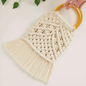 May include: Cream-colored macrame bag with a geometric pattern and a fringe detail at the bottom. The bag has two wooden handles. The bag is handmade and has a bohemian style.