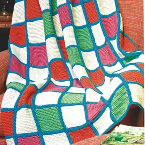 May include: A colorful knitted blanket with a patchwork design. The blanket features squares of red, pink, green, and white, outlined in blue. The blanket is draped over a red armchair, creating a cozy and inviting scene.