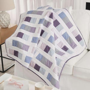 May include: A white quilt with rectangular patches of various fabrics in shades of purple, blue, and gray. The quilt is draped over a white sofa. The patches have striped, checkered, and solid patterns. The quilt has a dark purple border.