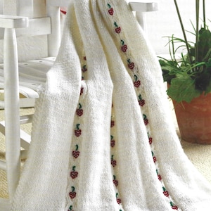 May include: A white knitted blanket draped over a white chair. The blanket features a repeating pattern of red berries with green leaves. The blanket is made of a soft, textured material and is suitable for home decor.