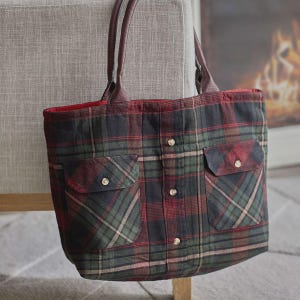 Könnte beinhalten: Eine karierte Tragetasche mit braunen Ledergriffen und rotem Innenfutter. Die Tasche hat zwei Vordertaschen mit Knopfverschlüssen und ist aus einem grün, rot und schwarz karierten Stoff gefertigt. Die Tasche steht auf einem grauen Stuhl.