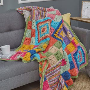 May include: A vibrant, hand-crocheted blanket with a patchwork design, draped over a grey sofa. The blanket features squares in various colours, including pink, blue, orange, and green. A white mug is on the sofa.