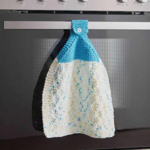 May include: A handmade, knitted kitchen towel with a button closure, hanging from an oven handle. The top portion is bright blue, while the main body is white with blue and yellow flecks. The towel is a practical and decorative kitchen accessory.