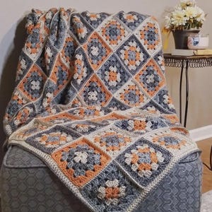 Peut inclure: Une couverture au crochet faite à la main, drapée sur un pouf gris. La couverture présente un motif répétitif de carrés dans des tons de gris, d'orange et de crème. La texture et les couleurs créent une ambiance chaleureuse et accueillante.