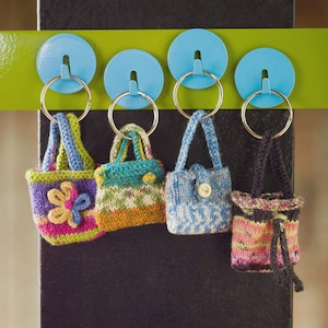 Può includere: Quattro mini borsette lavorate a mano a maglia in vari colori, ciascuna attaccata a un portachiavi argentato. Le borsette sono nelle tonalità del rosa, verde, blu e arancione, con motivi floreali e geometrici. Sono appese a ganci blu.