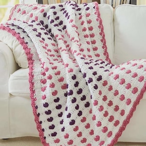May include: A white crochet blanket with a heart pattern in shades of pink and purple. The blanket has a pink border and is draped over a white sofa. This handmade blanket is perfect for home decor.
