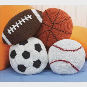 May include: A collection of handmade crochet pillows shaped like sports balls. Includes a brown football with white stitching, an orange basketball with black lines, a white baseball with red stitching, and a white and black soccer ball.