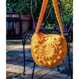 May include: A mustard yellow crochet shoulder bag with a circular design on the front. The bag has a long strap and is hanging on a black metal chair. The background includes a brick patio and greenery.