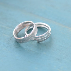 Op de afbeelding: Twee zilverkleurige ringen zijn in elkaar verstrengeld. De ene ring heeft een gladde, platte band met een subtiel, herhalend bloemmotief in het oppervlak gegraveerd. De tweede ring heeft een getextureerde, gehamerde afwerking met een open ontwerp.