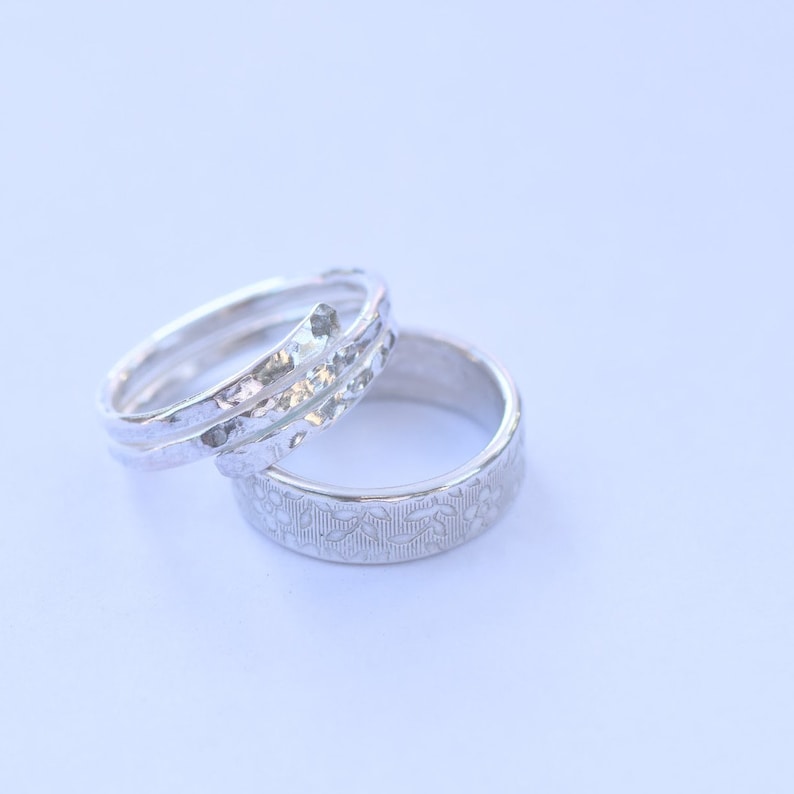 Op de afbeelding: Twee zilverkleurige ringen liggen bij elkaar. De ene ring heeft een overlappend ontwerp met een gehamerd, getextureerd oppervlak. De tweede ring is een platte band versierd met een herhalend, in reli&euml;f aangebracht bloemenpatroon.