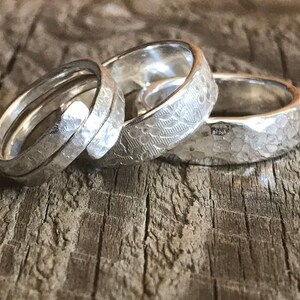 Op de afbeelding: Drie zilveren ringen op een rij. De eerste ring heeft een spiraalvormig ontwerp met een gehamerde textuur. De tweede ring toont een delicaat bloemenpatroon over het oppervlak. De derde ring heeft een opvallende, onregelmatige gehamerde afwerking.