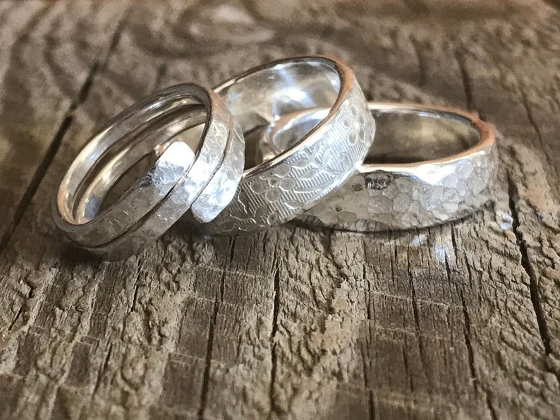 Op de afbeelding: Drie zilveren ringen op een rij. De eerste ring heeft een spiraalvormig ontwerp met een gehamerde textuur. De tweede ring toont een delicaat bloemenpatroon over het oppervlak. De derde ring heeft een opvallende, onregelmatige gehamerde afwerking.