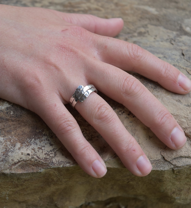 Op de afbeelding: Een zilveren ring met een bypass-ontwerp en een getextureerde, gehamerde afwerking. De band bestaat uit twee overlappende uiteinden die om de vinger wikkelen, wat zorgt voor een gelaagd uiterlijk met een metallic glans en onregelmatige oppervlakken.