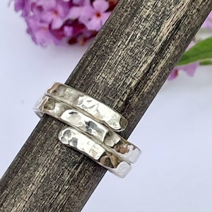 Op de afbeelding: Een zilveren ring met een gehamerde, getextureerde afwerking die om een donkere, verweerde houten stok is gewikkeld. De metalen band heeft een reflecterend oppervlak met meerdere inkepingen, wat een opvallend patroon over de gehele omtrek cre&euml;ert.