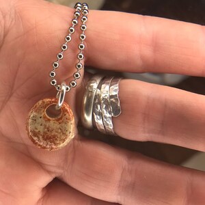 Op de afbeelding: Een zilveren kralenketting met een kleine, ronde, gespikkelde beige en roestkleurige keramische hanger, samen met een stapel van drie zilveren ringen, waarvan &eacute;&eacute;n met een gladde afwerking en twee met een gehamerd, getextureerd oppervlak.