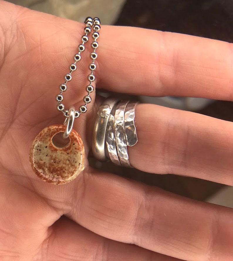 Op de afbeelding: Een zilveren kralenketting met een kleine, ronde, gespikkelde beige en roestkleurige keramische hanger, samen met een stapel van drie zilveren ringen, waarvan &eacute;&eacute;n met een gladde afwerking en twee met een gehamerd, getextureerd oppervlak.