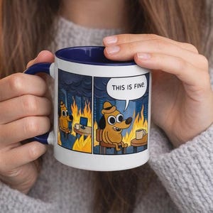 Könnte beinhalten: Weißer Keramikbecher mit blauem Innenraum und Henkel. Der Becher zeigt einen Cartoon-Hund in einem brennenden Raum, mit dem Text "THIS IS FINE" in einer Sprechblase. Ein humorvolles Design für Kaffee oder Tee.