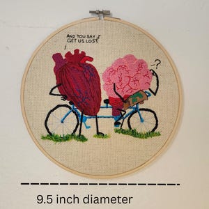 May include: Embroidered artwork on a 9.5-inch diameter hoop. The design features a heart and a brain riding a bicycle, with the text "AND YOU SAY / GET US LOST." The heart is red and the brain is pink.