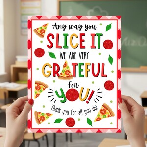 May include: A rectangular sign with a red and white checkered border, held in front of a classroom. The sign reads "Any way you slice it, we are very grateful for you!" with pizza slice and pepperoni illustrations. The background shows desks and a chalkboard.