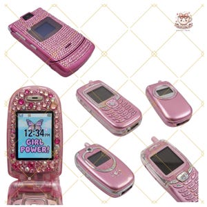 May include: A collection of pink flip phones, each adorned with rhinestones. One phone shows "GIRL POWER!" on the screen, with a butterfly and the time 12:34 PM. The phones have a glossy finish and different rhinestone patterns.