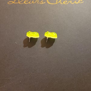May include: Two pairs of lime green gummy bear stud earrings. The earrings are on a dark brown surface with the gold text "Fleurs Cherie" in the upper portion of the image.