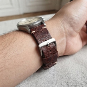 Pode incluir: Um relógio de pulso com uma caixa redonda de metal prateado e uma pulseira de couro castanho escuro texturizada. A pulseira é fixada com uma fivela de metal prateado e apresenta costuras claras visíveis ao longo das bordas da pulseira.