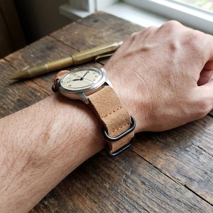 Peut inclure: Une montre-bracelet avec un boîtier rond argenté et un cadran crème doté de chiffres et d'aiguilles noirs. La montre est fixée par un bracelet en cuir suédé beige avec des coutures orange et deux passants métalliques noirs pour le réglage.