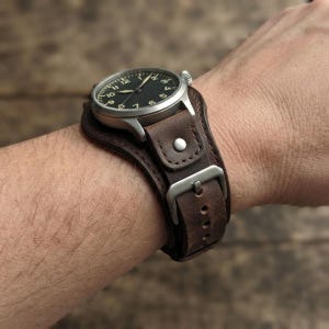 May include: A wristwatch featuring a round silver-tone case with a black dial and white numerals, attached to a wide brown leather cuff strap. The strap includes a metal buckle, a single metal rivet, and visible stitching along the edges.