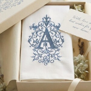 May include: A white fabric towel featuring a blue embroidered letter A surrounded by intricate scrollwork, presented in an open box with a matching lid, dried lavender sprigs, and a card with the text With love and best wishes for your new home.