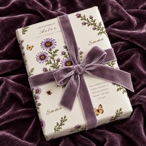 May include: A rectangular gift wrapped in cream-colored paper featuring purple aster flowers, butterflies, and a bee. The text reads September Aster, Love, Wisdom, Faith, and Emma. A wide, purple velvet ribbon is tied in a bow around the center.