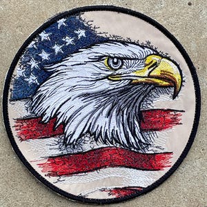 May include: Embroidered patch featuring an American bald eagle with a patriotic design. The patch showcases the eagle's head with white feathers, a yellow beak, and a background of the American flag in red, white, and blue.