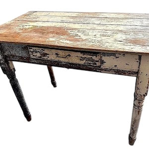 May include: A small, distressed wooden table with a single drawer. The tabletop has a weathered appearance with peeling paint and visible wood grain. The legs are turned and painted in a contrasting colour, adding to the antique aesthetic. The table is approximately 76 cm long.