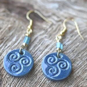 Könnte beinhalten: Ein Paar blaue Keramikohrringe mit goldfarbenen Haken. Jeder Ohrring hat ein kreisförmiges Design mit einem erhabenen Spiralmuster. Kleine blaue und goldfarbene Perlen sind am Haken befestigt.