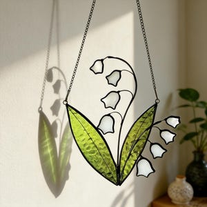 May include: A stained glass lily of the valley hanging decoration. The piece features white bell-shaped flowers and green leaves, outlined in black. It hangs from a chain, casting a shadow on the wall. The design is reminiscent of spring and nature.