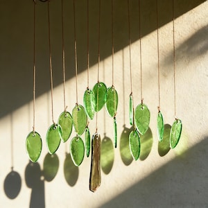 May include: A handcrafted wind chime featuring multiple green glass leaf shapes suspended by thin, brown cords. The leaves vary in size and shape, with a few rectangular glass pieces. The chime hangs against a neutral wall, with sunlight casting shadows.