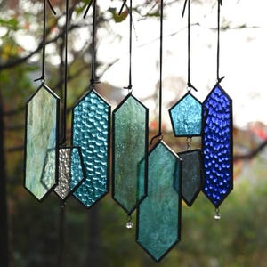 Peut inclure: Une collection de capteurs solaires en vitrail de différentes formes et nuances de bleu, vert et argent. Chaque pièce est bordée de noir et suspendue à un cordon noir. Certains ont du verre texturé et des accents de cristal.