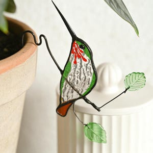 May include: A stained glass hummingbird decoration with a long black beak, red and green accents, and silver-coloured body. The bird is perched on a black wire branch with two green glass leaves. The hummingbird is designed to hang on a plant pot.