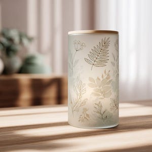 May include: A cylindrical glass candle holder with a wooden top. The frosted glass is decorated with delicate botanical illustrations in shades of green and brown. The candle holder sits on a wooden surface, with soft light illuminating the design.