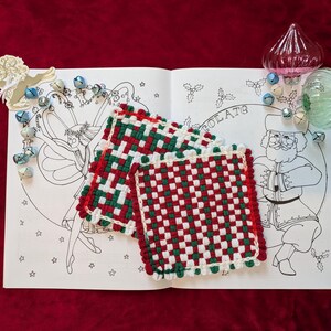 May include: Two red, green, and white woven square pot holders rest on an open coloring book with a Santa Claus illustration. Decorative glass ornaments and small bells are placed around the book. The background is a deep red fabric.