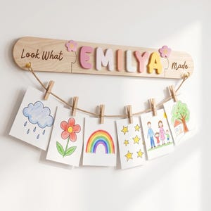 May include: A light wood wall display board with the text Look What EMILYA made in colourful 3D letters and two pink flower accents. A jute rope hangs from the board with six wooden clothespins holding children's drawings of a cloud, flower, rainbow, stars, family, and house.