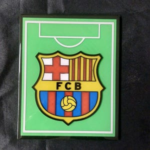 Może przedstawiać: Prostokątny zielony znak z herbem FC Barcelona. Herb ma kształt tarczy z czerwonym krzyżem, pionowymi paskami i piłką nożną. Litery "FCB" znajdują się na dole.