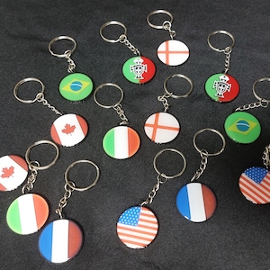 May include: A collection of keyrings with circular pendants displaying national flags, such as the USA, Brazil, Canada, France, Italy, Portugal, and England. Each keyring has a silver chain and ring.